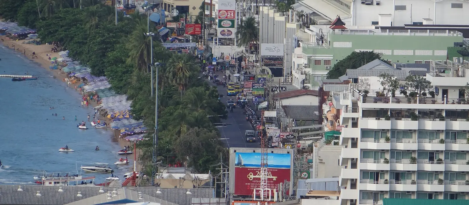 Pattaya Beach Road seen from above: the beach and its palms on the left, hotels, shops and traffic along the road on the right.