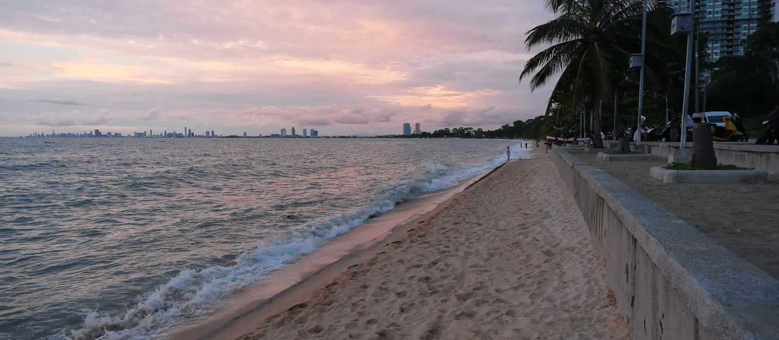 A quiet sandy beach and sea wall at sunset, with high-rise towers in the distance.